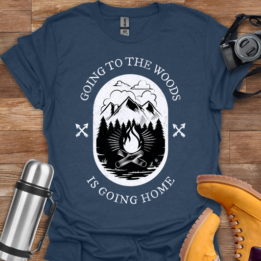 A navy blue crew neck T-shirt with a graphic print of mountains and text 'GOING TO THE WOODS IS GOING HOME' displayed on it, presented flat on a wooden surface alongside a thermos and a pair of boots.